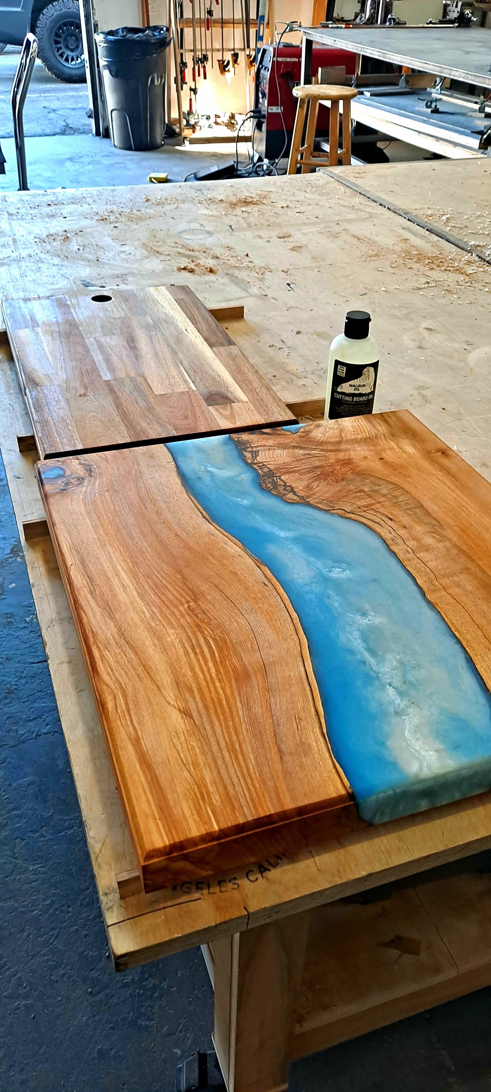 Live-edge slab with a poured blue epoxy river resting on the workbench next to a bottle of finish