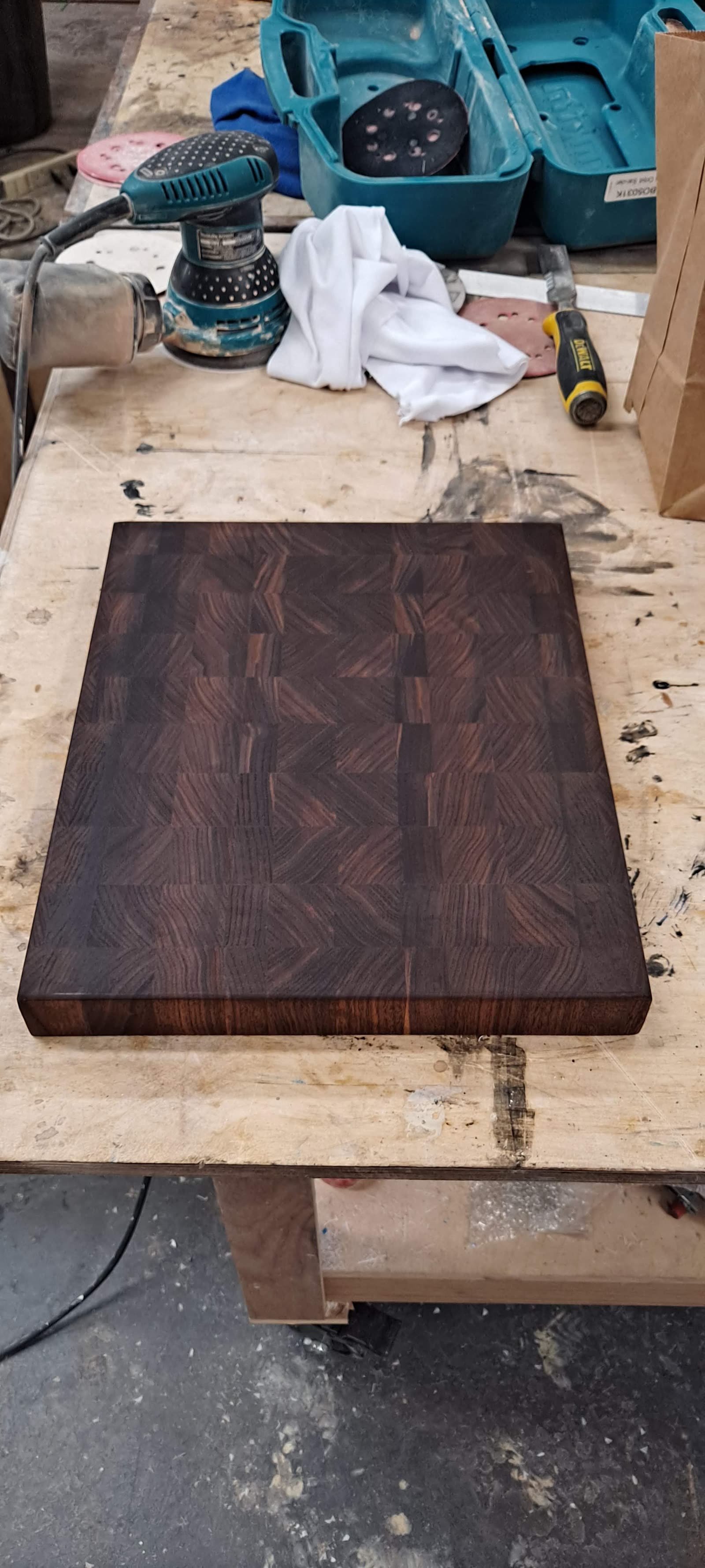 Thick dark end-grain walnut cutting board sanded and sitting on the workbench beside an orbital sander