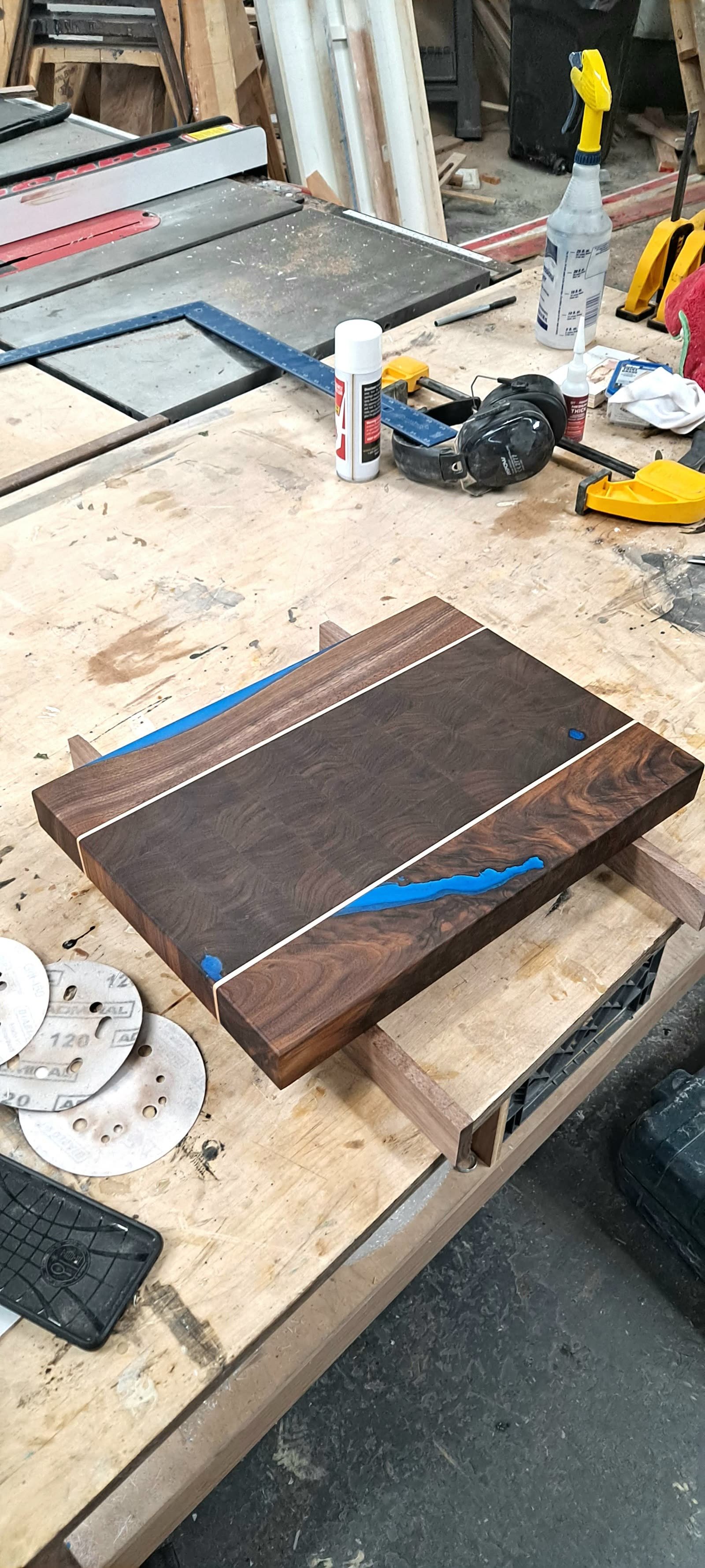Dark walnut end-grain cutting board with pale accent stripes and blue epoxy inlays, resting on risers in the shop
