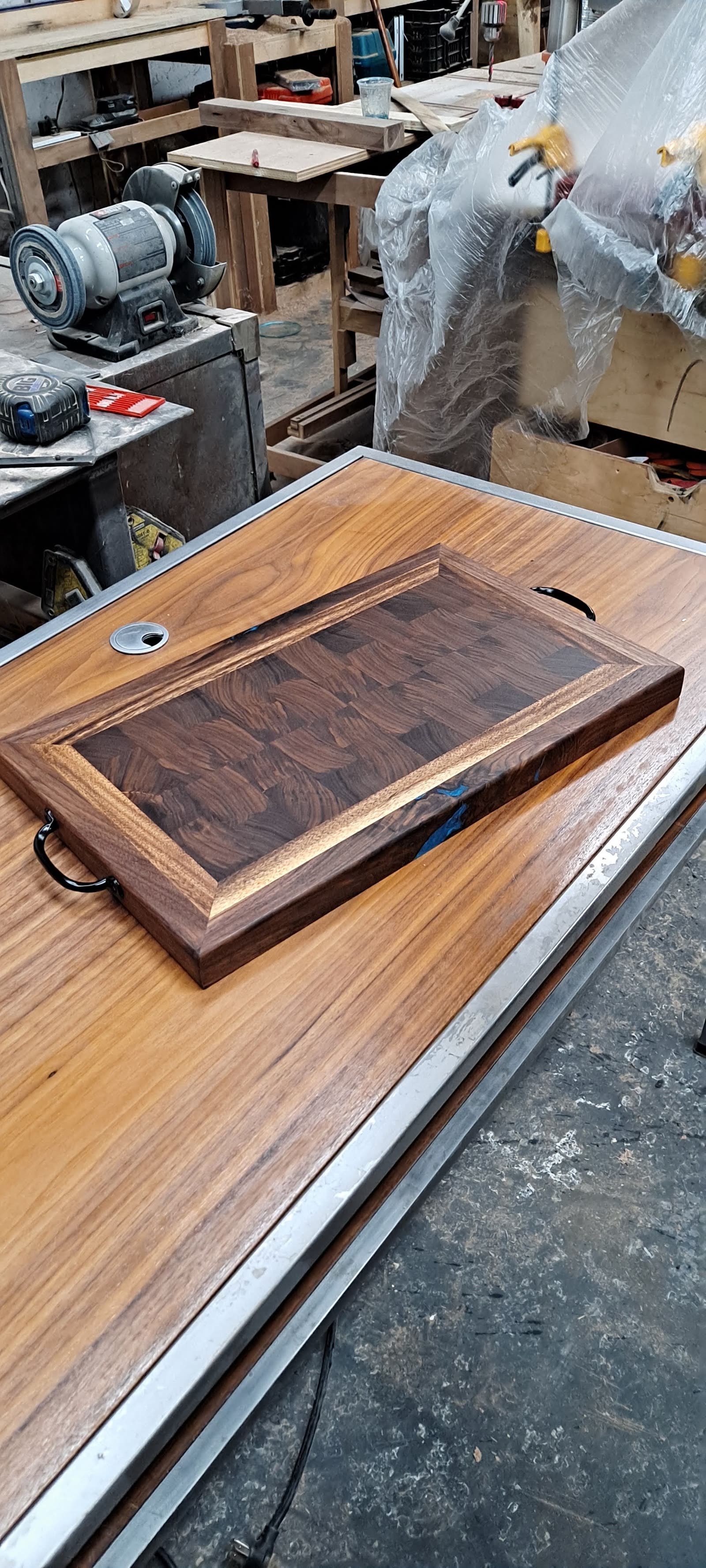 End-grain carving board with a routed juice groove and cut-in handles, resting on a wide hardwood tabletop in the shop